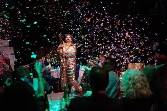 A man in a gold boiler suit stands in the middle of tables and people, releasing a confetti cannon into the air.