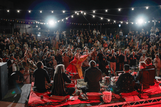 Two bright lights overlook a crowd of people sat in a roman-style amphitheatre, with a group of performers sat on the stage in the foreground.