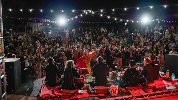 Two bright lights overlook a crowd of people sat in a roman-style amphitheatre, with a group of performers sat on the stage in the foreground.