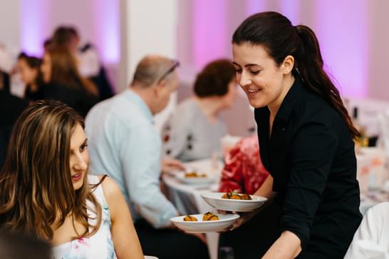 In the background, there are tables with people enjoying meals. In the foreground a woman to the left is seated smiling, and to the left a waitress brings plates of food to the table smiling.