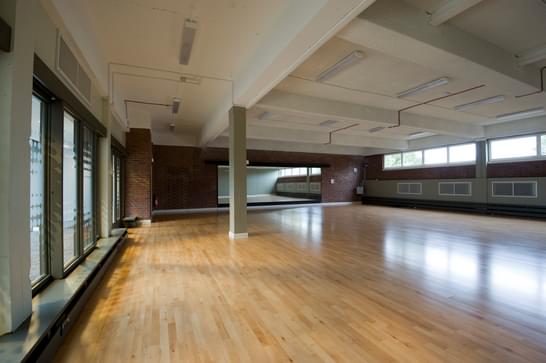 A large room with a long mirror alongside the far wall, with floor-to-ceiling windows that lets the light flood in across the hardwood floors.