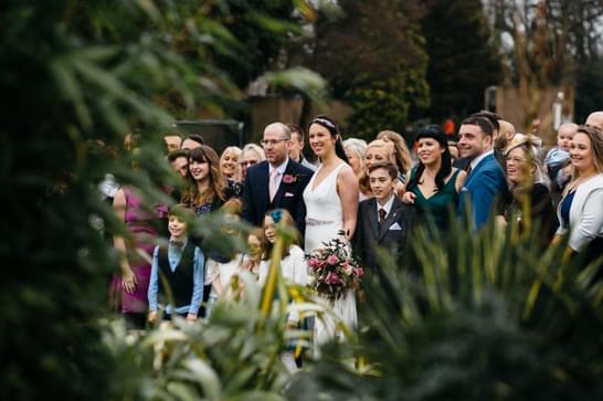 Leaves surround the outer edge of the image, and a wedding party can be seen through the middle. A bride and groom stand surrounded by their wedding party.
