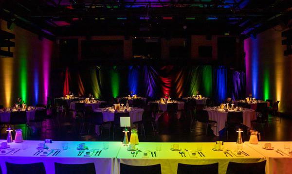 A wide shot of a theatre, dressed for a wedding. To the bottom, there is the head table and in the background there are many tables surrounded by multicoloured lights