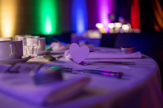 A close up of a table with a a white table cloth, and a white paper cut out heart. In the background, the room is lit up with rainbow colours.