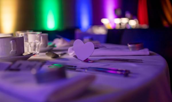 A close up of a table with a a white table cloth, and a white paper cut out heart. In the background, the room is lit up with rainbow colours.