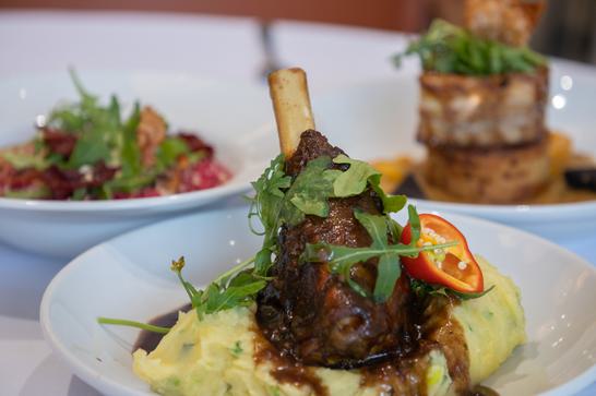 At the centre of the dish, a leg of lamb is sitting on some mashed potato and garnished with salad and jus. In the background, two other well crafted dishes are slightly blurred.
