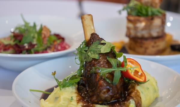 At the centre of the dish, a leg of lamb is sitting on some mashed potato and garnished with salad and jus. In the background, two other well crafted dishes are slightly blurred.