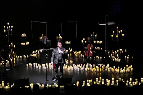 A man is in the middle of a large, dark stage and is surrounded by hundreds of candles.
