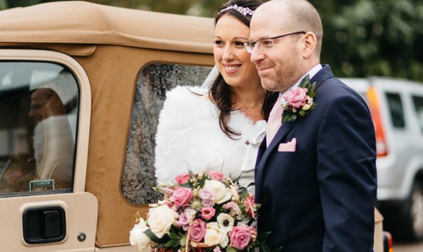 A bride and groom are photographed outside next to an old style car. They are smiling and linked by their arms.