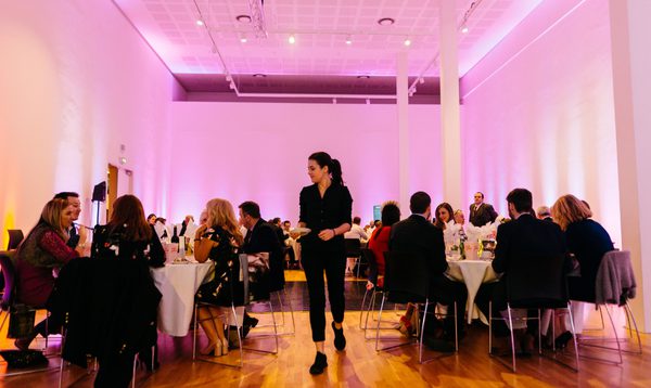 A wide shot of a wedding room, filled with people sitting and white clothed tables. A servers walks through the middle with a plate of food.