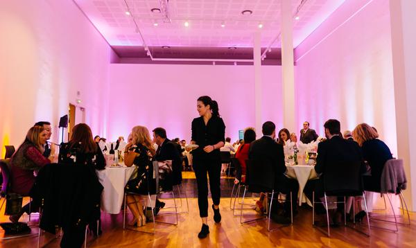 A wide shot of a wedding room, filled with people sitting and white clothed tables. A servers walks through the middle with a plate of food.