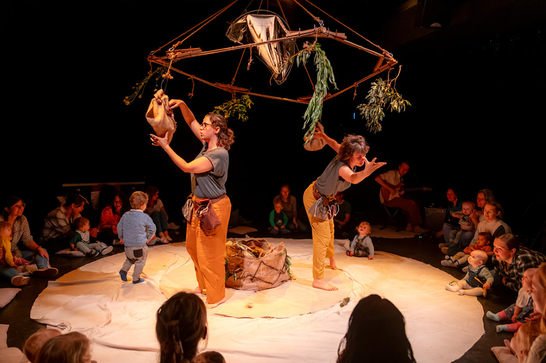 Two performances stand underneath a giant mobile ornament on stage which has various objects hanging from it - they are surrounded in a circle by an audience of babies and adults.