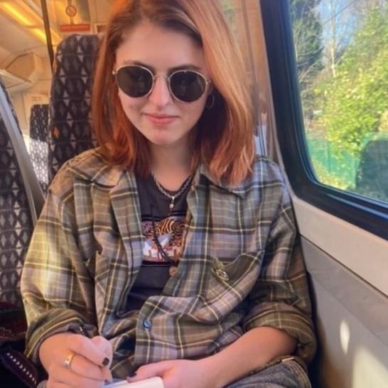 Georgia is seated on a train, and is wearing a checked shirt and sunglasses. Georgia has long, brown hair.