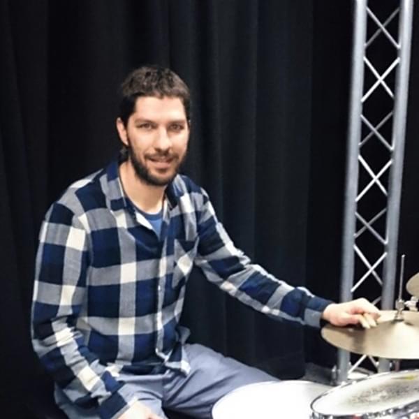 Ilias has short brown hair and a short brown beard. Ilias is sitting at a drum kit.