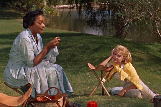 Two women (Lana Turner and Juanita Moore) sit in a sunny park, one resting her arm on a stool.