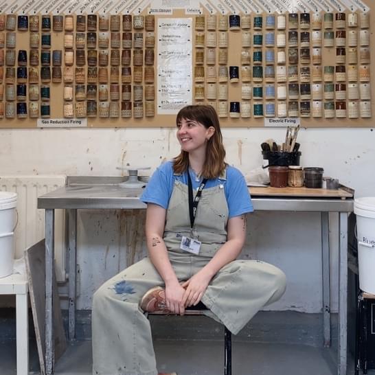 Imogen is sitting with one leg crossed in a ceramic studio, wearing grey dungarees. Imogen has medium length brown hair and a fringe.