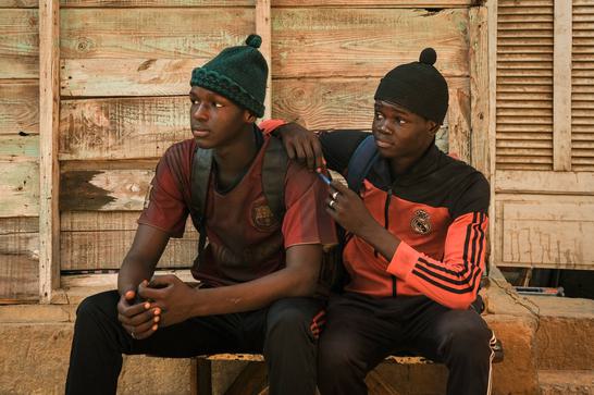 Two Senegalese teenagers wearing bobble hats and wearing backpacks sit on a bench, looking off to the left.