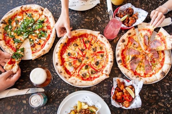 Birds-eye view of pizzas and drinks, people's hands grabbing the food at a table