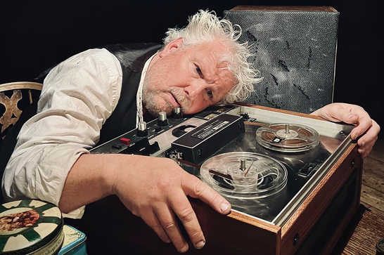 David Westhead is sitting down and resting his head on top of a large tape player with his hands on either side of it.