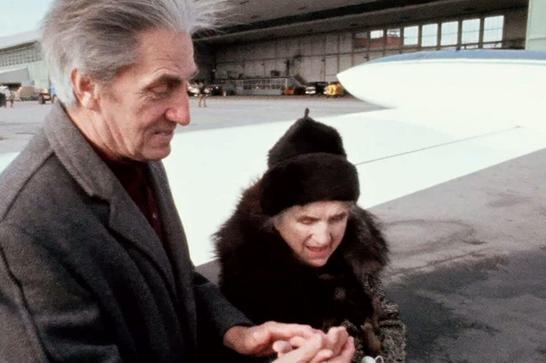 A deaf-blind man and a deaf-blind woman hold hands as they exit a small airplane.