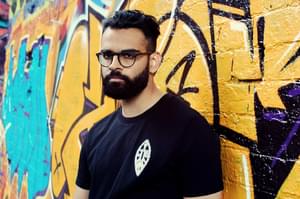 A man with dark brown hair and a beard, wearing glasses and a black tshirt