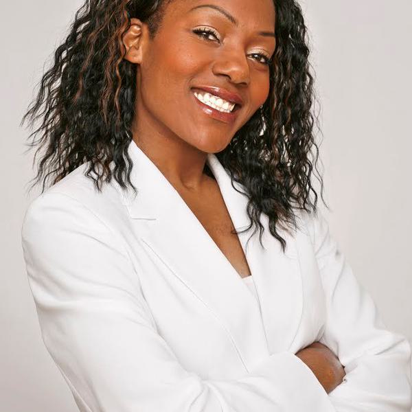 Nataylia has dark brown hair, is wearing a white suit, and is stood with her arms crossed and smiling