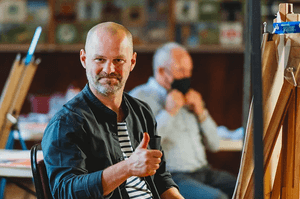 Nick sits at a wooden easel, wearing a green shirt and is giving a thumbs up to camera