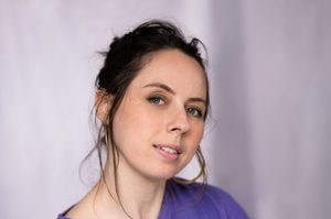 A woman is wearing a purple top, with brown hair tied up with a few strands falling around her face.