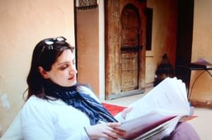 Unaiza Karim is sitting in a chair, reading a book. Unaiza has long brown hair and has glasses on top of their head.