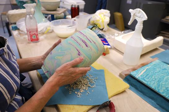 A person has their hands outstretched, holding a marbled coloured vase with both hands.
