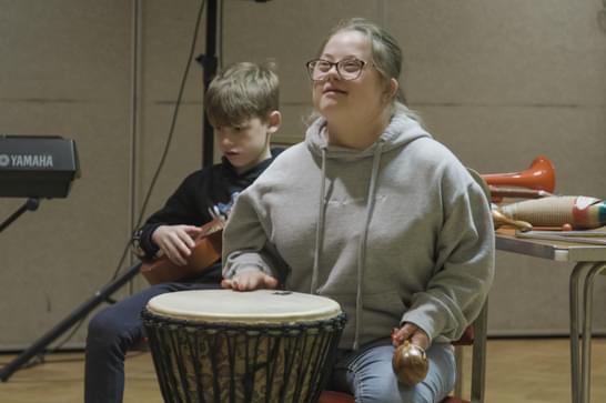 A young girl in a grey jumper bangs a drum with her hand.