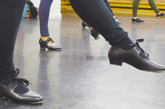 outstretched legs with tap shoes on, in a dance studio with mirrored walls behind them