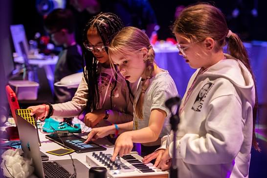 thee young girls stand at music making table, featuring keyboards and mixing boards. The background is lit up with purple light.