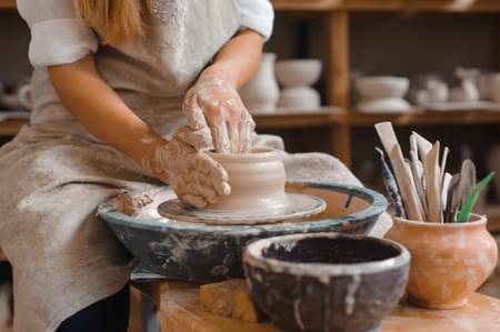 A woman is sitting at a pottery wheel, sculpting a pot with her hands. To the left, there are sculpting tools next to her.