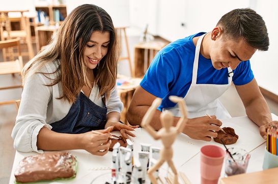 A couple are sat together at a table wearing aprons, both are using their hands to mould clay