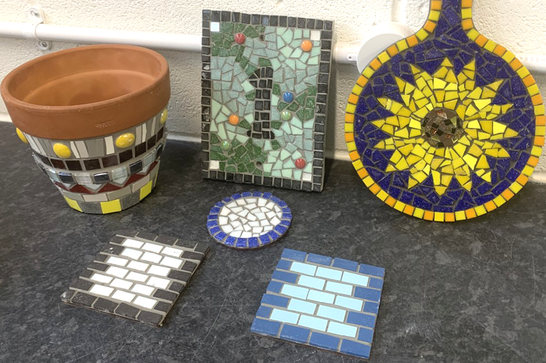 A collection of items - including coasters, a terracotta pot and a plaque - decorated with brightly coloured mosaic tiles.