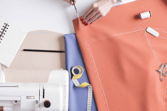 A person is cutting fabric, with drawn outlines on it. There is measuring tape, thread and a sewing machine to the sides.