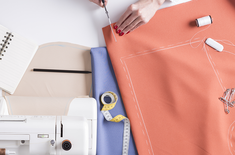A person is cutting fabric, with drawn outlines on it. There is measuring tape, thread and a sewing machine to the sides.