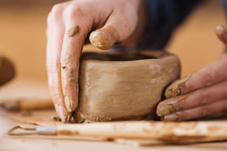 A person is moulding a block of clay with their hands, with tools to the front left