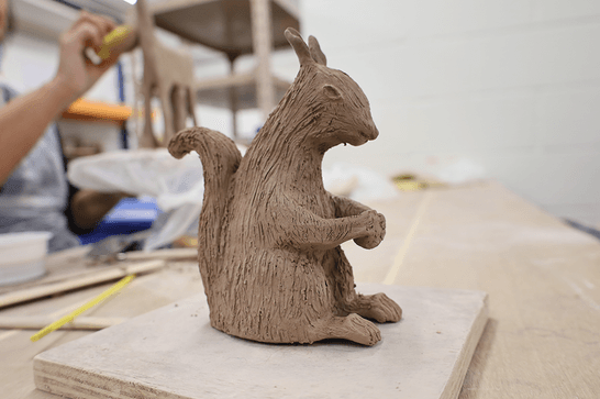 A clay squirrel is sitting on a board, in the background someone uses a sponge to smooth a clay giraffe