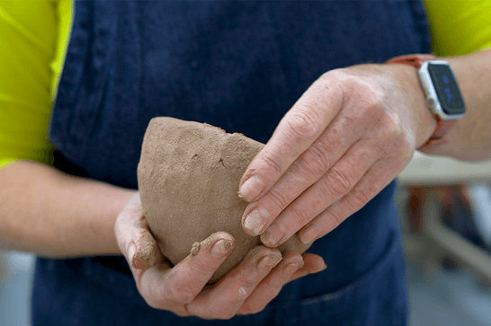 A person in a green top and blue apron is holding a clay bowl with both hands