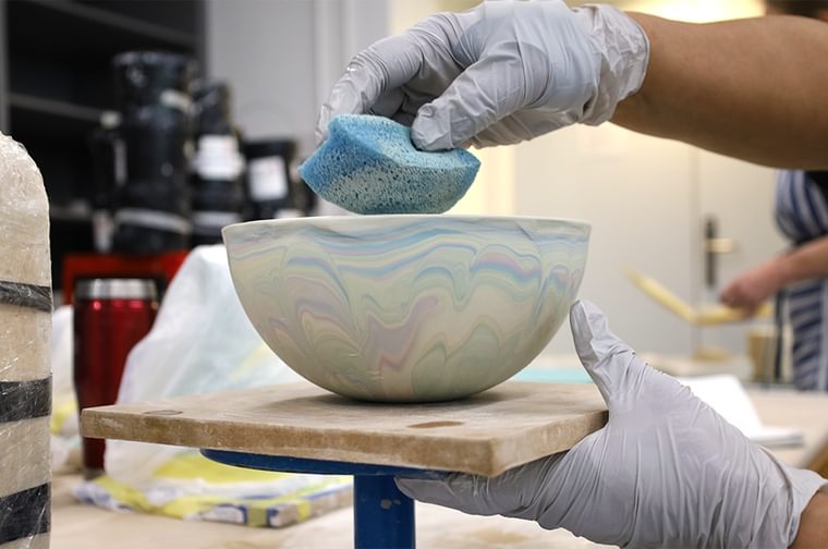 A person is using a sponge to smooth down a blue and purple marbled bowl