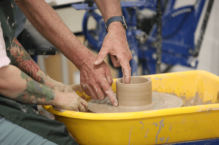 A person's hands are forming clay on a pottery wheel, with a second pair of hands assisting