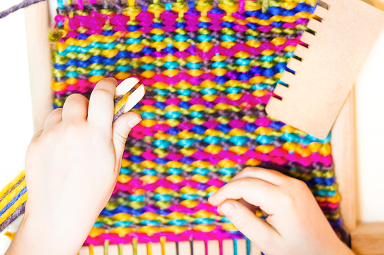A young child has a loom in front of their hands, with colourful wool weaved onto it