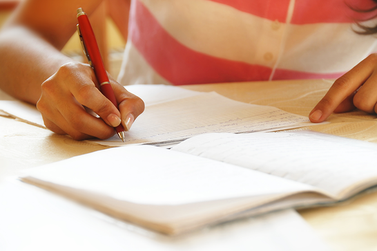 A person is sat at a desk, with a pink and white stripey top, and is writing on a blank piece of white paper