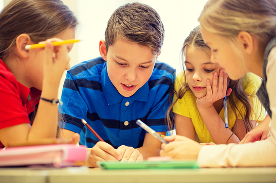 Four children are gathered around a table holding pens, all looking at a piece of writing