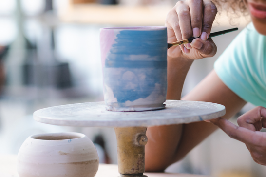A pot is being painted with a small paintbrush, in blue and pink paints