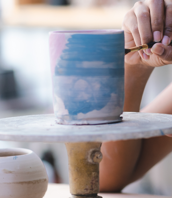 A pot is being painted with a small paintbrush, in blue and pink paints