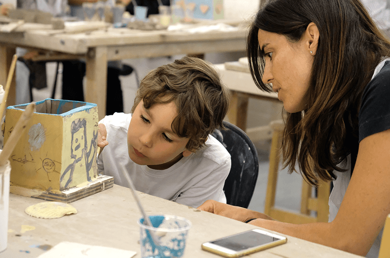 A young child is painting a clay box, with a woman leaning over him to see what he is doing