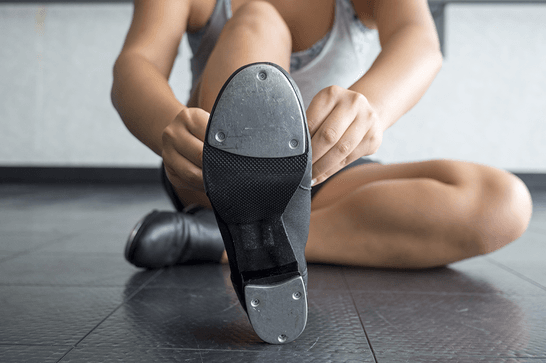 A person is sat with one leg tucked below them, putting on a tap dance shoe